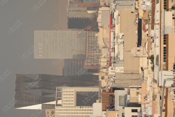 A cityscape featuring modern skyscrapers, including a tall building with "KAFD" written on it. The buildings display various shades of beige and brown, with a few cranes visible amidst construction. The sky is clear with daytime lighting.