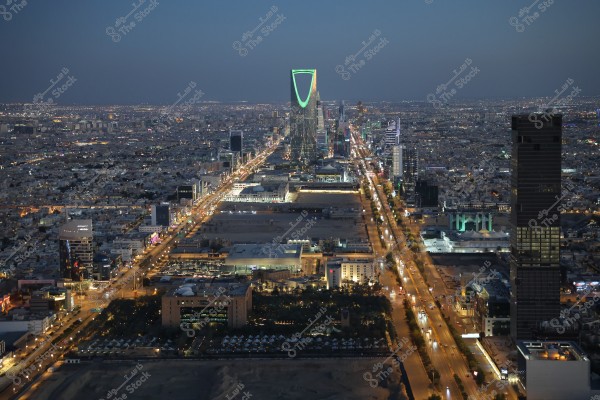 منظر جوي لمدينة الرياض في المملكة العربية السعودية ليلًا, حيث يظهر برج المملكة مضاءً بألوان زاهية, ويقف وسط المدينة. تمتد الشوارع المزدحمة بالإضاءة والنشاط على كلا الجانبين, مع وجود العديد من المباني الشاهقة التي تشكل أفق المدينة.