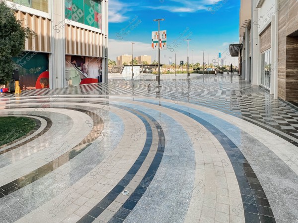An image depicting an outdoor plaza in a modern commercial area, featuring a multicolored tile flooring with geometric patterns. The surrounding buildings have glass facades and artistic decorations. The sky is blue with some clouds, and greenery adds a natural touch to the scene.