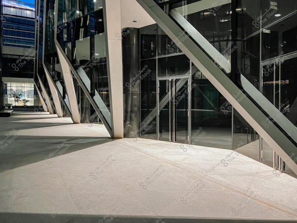 The image shows a section of a modern building with a steel and glass structure. The architecture features slanted supports and reflective glass extending across the facade. The surrounding area appears clean and is illuminated with soft lighting, highlighting the contemporary architectural design.
