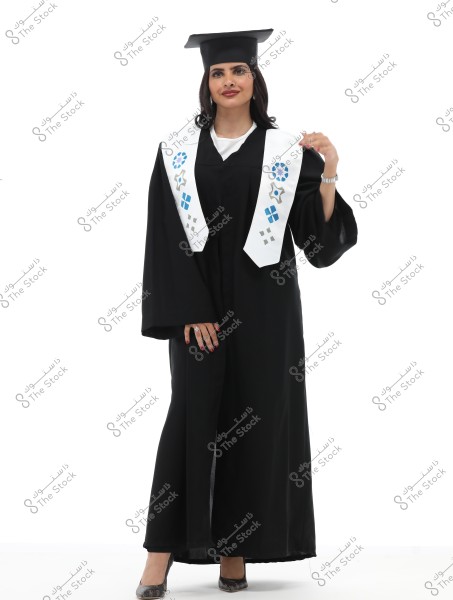 An image of a woman wearing graduation attire consisting of a black gown and a white graduation stole adorned with blue and silver geometric patterns. She is wearing a black graduation cap and stands smiling, conveying a sense of pride and achievement. Her shoes are black with decorative patterns, and the background is completely white.
