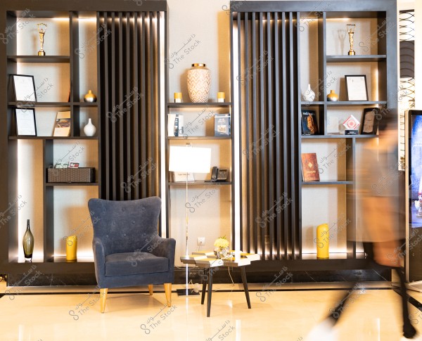 The image shows an interior decor corner with shelves holding trophies, frames, pictures, and books. A comfortable blue chair sits in the center, accompanied by a small table with some flowers. A blurred person is moving on the right side of the image. The lighting is warm, and the geometric lines are elegant.