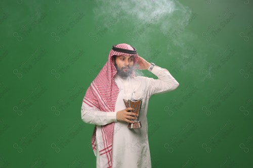 A Saudi man standing on a green background wearing a thobe and a shemagh, making gestures with his hands, steaming himself with perfume and agarwood, Eid celebrations