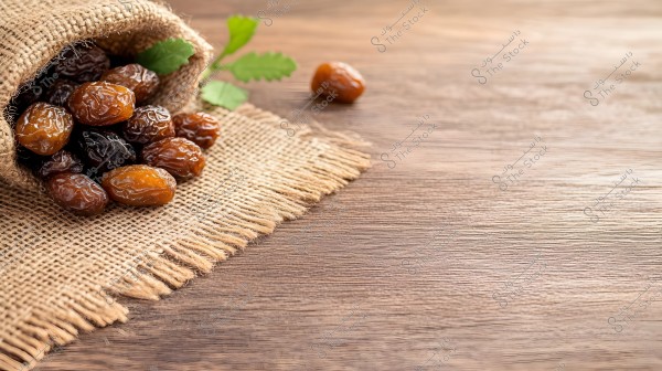 An image showing several dates scattered on a piece of burlap fabric. The burlap is light brown and rests on a wooden table, with some fibers hanging off. A small green leaf is visible next to the dates on the fabric.