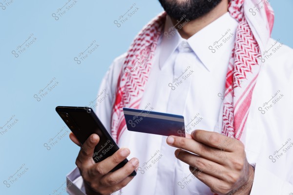 Image of a man wearing traditional Saudi attire, consisting of a white thobe and a red and white ghutra. He is holding a smartphone in one hand and a bank card in the other. The background is light blue.