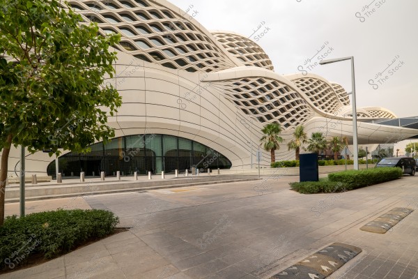 A modern architectural building with curved surfaces and large windows, featuring a lattice pattern on the roof. There are palm trees in the foreground, along with other small trees and shrubs lining the sidewalk. The road and pavement appear clean and clear of pedestrians.