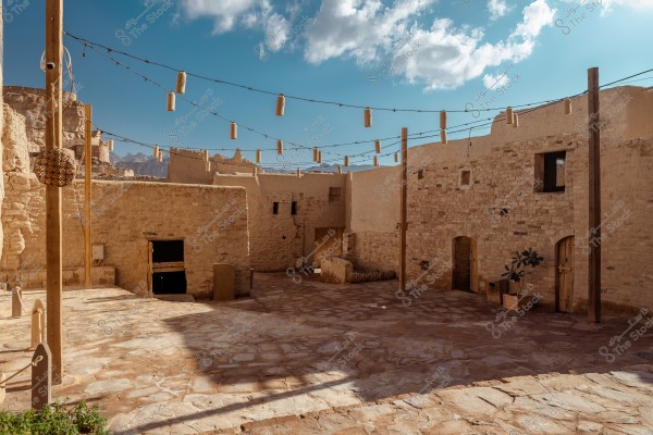 An inner courtyard in a traditional historical building, featuring stone and mud walls, with wooden doors and string lights hanging between wooden posts. The sky is clear with a few scattered clouds.