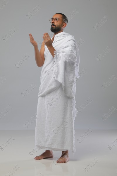 A portrait of a man in a prayer position wearing traditional white Ihram clothing, consisting of two pieces of unstitched white fabric. The man is standing on a smooth white floor, with his hands raised in prayer and wearing round glasses. The background is light grey.