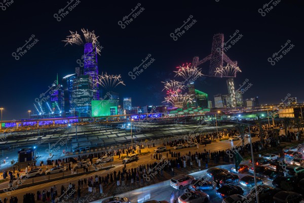 A nighttime view of a modern city skyline illuminated with vibrant colors and fireworks lighting up the sky. The skyscrapers are lit in hues of blue, green, and purple. People are gathered below along the road watching the display, highlighting the unique architectural style with lights and firework effects. Cars are parked along the sides of the road.