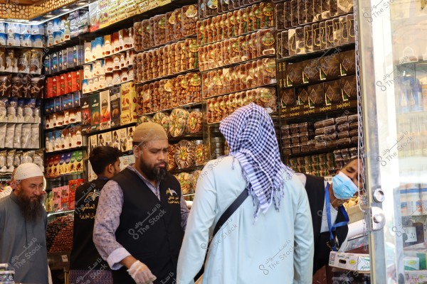 The image shows a shop filled with food products, with the upper walls displaying various colorful and packaged items. Several men are in the store wearing traditional clothing; one is in a white thobe and a traditional checkered headscarf, another in a black vest with Arabic writing. One person is wearing a face mask and is focused on arranging products.