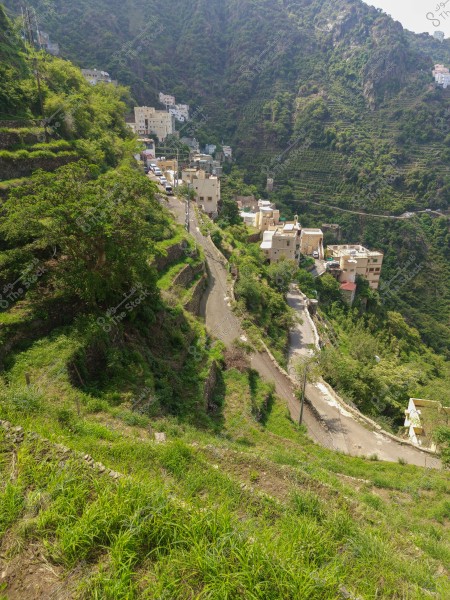 A scenic view of a mountainous village with lush green terraced terrain. Traditional-style homes are visible on the mountain slopes, surrounded by trees and greenery. A winding road runs through the village, adding a peaceful and picturesque quality to the area.