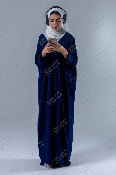 صورة لامرأة ترتدي عباءة زرقاء وحجاب أبيض, ترتدي سماعات رأس سوداء وتنظر إلى هاتف محمول. تبدو وكأنها في وسط نشاط يومي بسيط. تتناسب الإضاءة الرمادية الخلفية بشكل جيد مع ملابسها.
