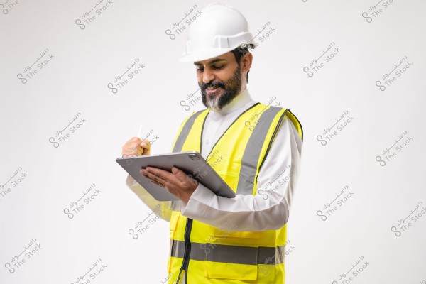 Image of a man wearing a white hard hat and a yellow safety vest over a white thobe, suggesting a Gulf region origin like Saudi Arabia. He is holding a tablet and writing on it with a stylus. The background is plain white.