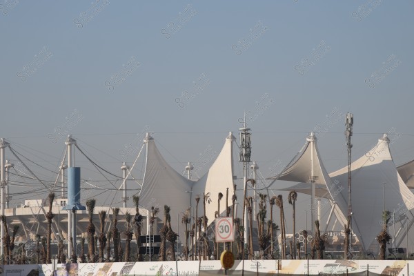 The image shows large white structures resembling tents, which are part of a modern architectural design. In front, there are several palm trees and some metal towers, along with a sign indicating a speed limit of 30 km/h. The sky is clear, suggesting an urban setting.