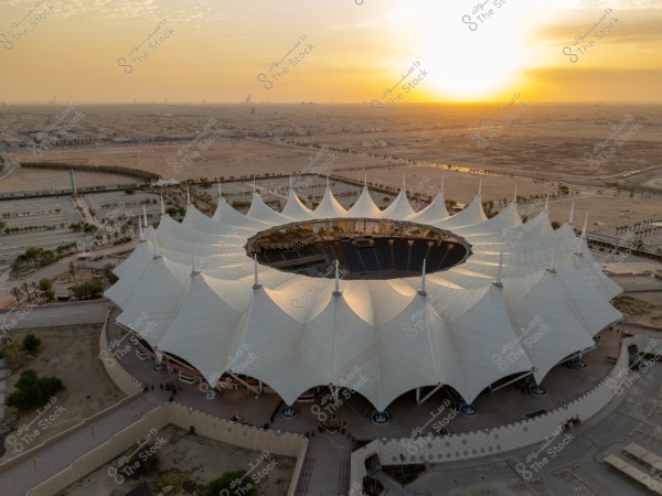 The image shows a large sports stadium with a white fabric roof designed in a striking geometric pattern resembling tents, seen from an aerial view against a desert city backdrop at sunset. The sky is clear with a warm orange hue as the sun sets on the horizon, and the stadium is surrounded by open spaces, some trees, and sandy areas.