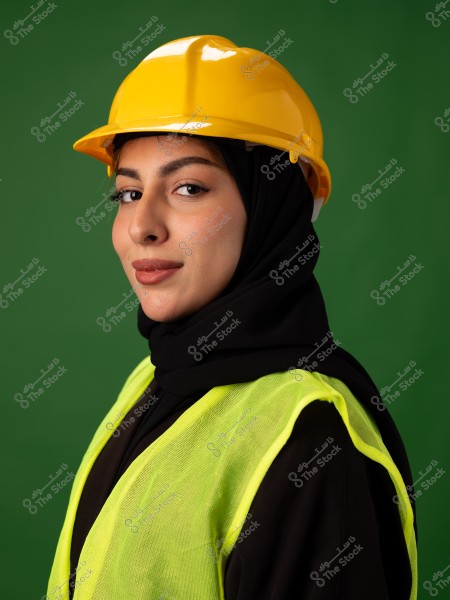 Portrait of a woman wearing a yellow safety helmet and a green reflective vest, with a black hijab. The background is green, highlighting her confident facial features. She appears to work in an environment that requires safety gear.