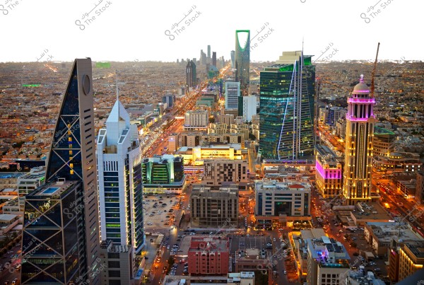 Aerial view of Riyadh, Saudi Arabia, featuring numerous modern skyscrapers and tall buildings. The iconic Kingdom Tower is visible prominently in the background, surrounded by buildings with varied architectural designs. The streets are illuminated and appear busy with traffic, while the buildings are lit with vibrant colors.
