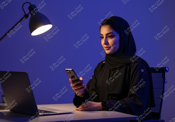 صورة لامرأة ترتدي الحجاب والعباية السوداء تجلس على مكتب مضاء بمصباح مكتبي. المرأة تحمل هاتفًا ذكيًا أمامها وهناك جهاز لابتوب على المكتب. الأجواء زرقاء داكنة توحي بالهدوء والتركيز.