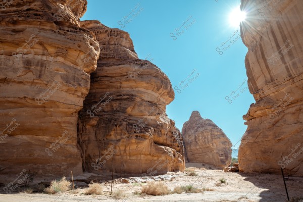 Image shows massive rock formations in a desert area under a clear blue sky, with the sun shining in the upper right corner. The rocks have layers of brown and beige colors, with shadows casting on the sandy ground and small dry plants.