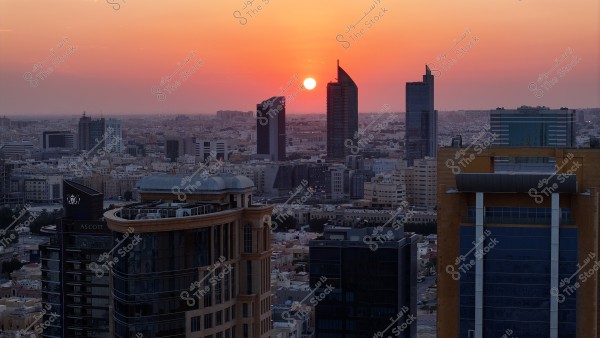A photo of a modern city at sunset, with skies glowing in warm orange hues. In the foreground are several tall buildings, some with sleek, modern glass designs, and others more traditional. The sun is setting behind a modern tower on the horizon, casting a mix of shadows and lights across the skyline.