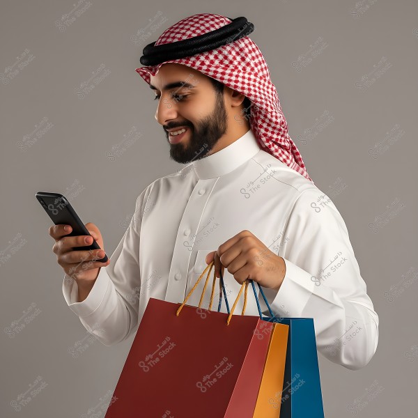 Image of a man wearing a white thobe and a red checked headscarf with a black agal, holding two colorful shopping bags and looking at his phone with a smile. The background is neutral gray, and the attire suggests traditional clothing from the Gulf region.