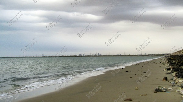 A sandy beach under a cloudy sky, with gentle waves lapping at the shore. A series of tall buildings can be seen on the horizon, indicating a distant city. Rocks are scattered along the beach, and the overall atmosphere appears calm yet slightly gloomy due to the heavy clouds.