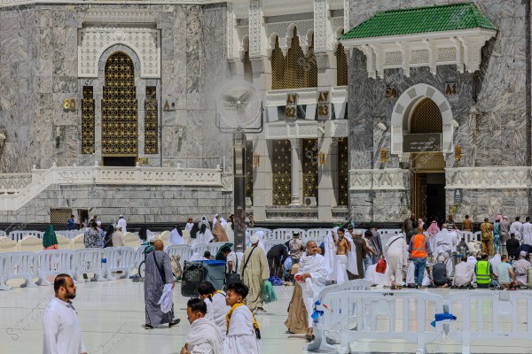 A group of people walking and moving in the courtyard of the Masjid al-Haram in Mecca, Saudi Arabia. In the background, the mosque\'s walls are adorned with marble and Islamic architectural decorations. Some individuals are wearing traditional white Ihram garments, while others are in casual attire. The scene depicts daily life and movement near the mosque.