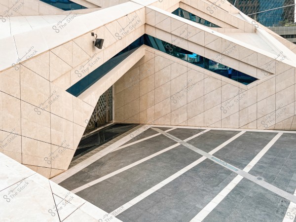 An image of a modern architectural facade with a distinctive geometric design. The walls are covered with white tiles featuring defined lines, and the slanted glass windows add a contemporary touch. In front of the building is a marble floor with intersecting lines of contrasting colors. A metal door is visible at the corner of the building.
