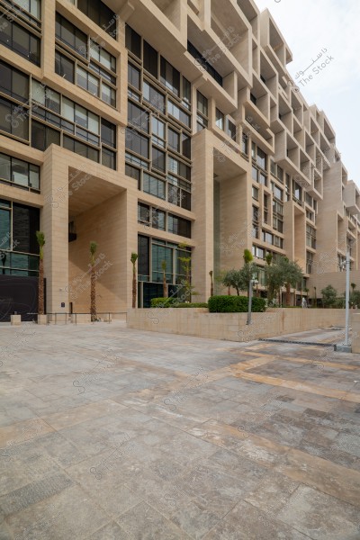 The image shows a modern multi-story building with contemporary architectural design. The facade features large glass windows and beige stone frames. In front of the building, there is a paved plaza with gray stones, and some trees and plants are arranged along the frontage.