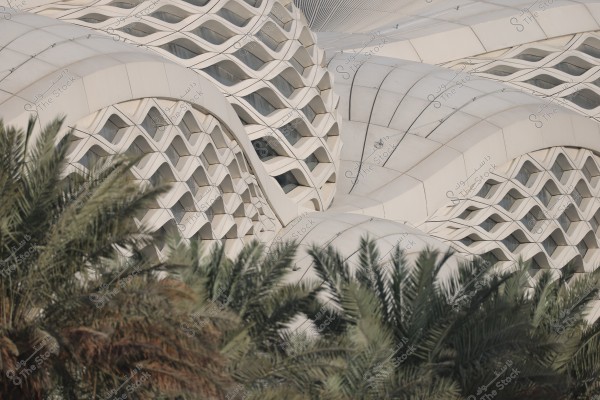 The image shows a modern architectural design with intricate geometric lattice patterns on the building\'s facade. In the foreground, there are green palm trees adding a natural element to the scene. The design reflects artistic interlocking and precision in details.