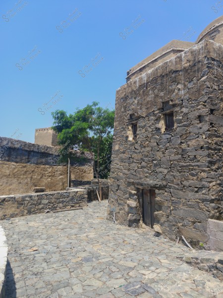 An image of an old stone building on a sunny day, characterized by thick walls made of large rocks. The stone-paved path is visible in front of the building, while green trees stand beside it, with a clear blue sky in the background. The architectural style suggests traditional Middle Eastern design.