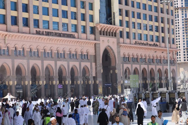 A large group of people walking in front of the \"Dar Al Tawhid Intercontinental\" hotel in Mecca, Saudi Arabia. The building features Islamic architectural design with arches and traditional decorations. Many people are wearing the traditional white clothing of ihram, indicating they may be pilgrims or performing Umrah. There are advertisements and offices on the ground floor of the building.