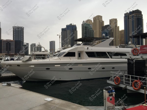 Image of a luxury white yacht docked at a marina with a backdrop of tall buildings. The scene includes several modern skyscrapers under a cloudy sky. There is a pier on the side with orange life buoys.