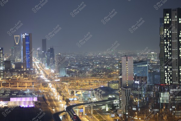 A nighttime cityscape of Riyadh, showcasing tall skyscrapers and modern buildings, including the iconic Kingdom Tower with its distinctive spire shape. The city is illuminated beautifully, with lights spread along the highways surrounding the structures, highlighting the modern architectural design of the city\'s infrastructure.