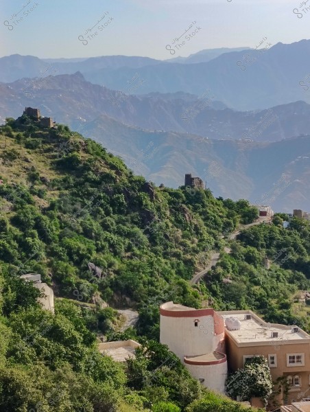 A scenic view of a mountain covered in greenery and buildings overlooking a valley in a mountainous region. The image shows hills and mountains in the background with a clear blue sky.