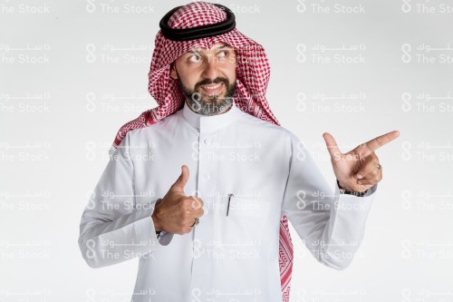 بورتريه رجل سعودي يقف على خلفية بيضاء، يرتدي الثوب، ينظر الى الكاميرا وهو مبتسم ويقوم بعمل حركات بيديه