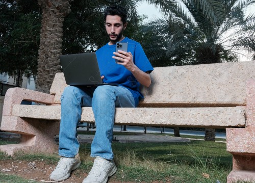 A Saudi man sits on a sofa in a public park and uses a mobile device during the day.