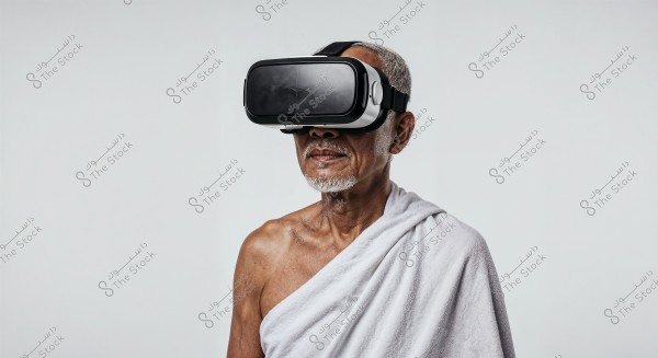 An image of an elderly man wearing black virtual reality goggles, dressed in a white cloth wrap resembling Ihram clothing, covering his shoulder. The background is plain white with no details.