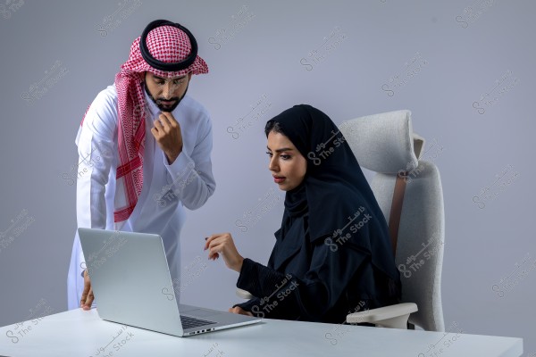 The image depicts a man and a woman at a desk reviewing content on a laptop. The man is standing and is dressed in traditional Saudi attire, including a thobe, shemagh, and agal. The woman is seated on a chair, wearing a black abaya and hijab. The room is neutral in color, highlighting the focus on the interaction between the two individuals.