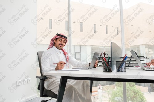 رجل سعودي يرتدي الزي السعودي التقليدي يعمل في مكتب بواجهة زجاجية ويستخدم قلم رصاص لتدوين الملاحظات