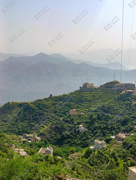The image depicts a mountainous landscape with green terraces spread across its slopes, where buildings are scattered sparsely. A range of mountains can be seen in the background under a hazy sky. The features suggest a rural area, likely in Saudi Arabia.