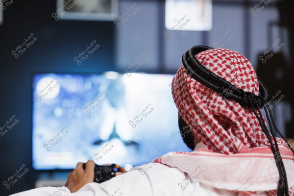Image of a man wearing a thobe, ghutra, and agal, sitting in a somewhat dimly lit room in front of a large TV screen with a blue glow. He appears to be playing a video game using a controller. The traditional clothing suggests he might be from Saudi Arabia or the Gulf region.