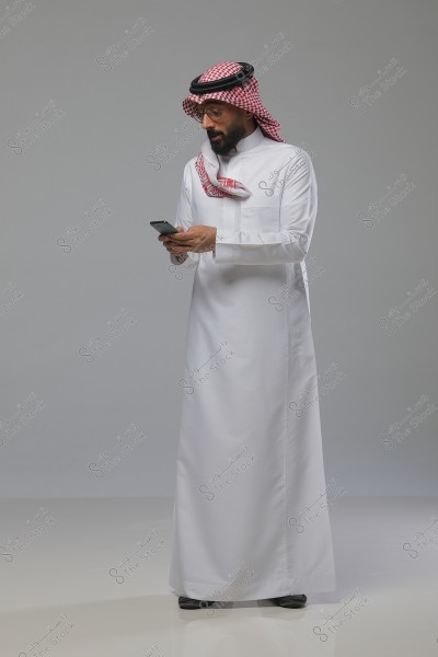 صورة لرجل يرتدي ثوبًا أبيض تقليديًا وغترة حمراء مع عقال أسود. يحتضن بيده هاتفًا محمولًا وينظر إليه بانتباه. الخلفية رمادية وملامح الصورة تعكس الوضعية الوقوف.