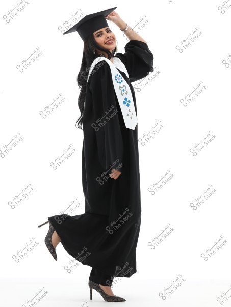 An image of a woman wearing a black graduation gown and matching graduation cap. She wears a white graduation stole adorned with colorful decorations. The woman is smiling and lifting her arm to touch the cap. She is wearing patterned high-heeled shoes, and the background is white.