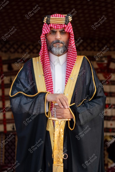 A portrait of a man wearing traditional Saudi attire. He is dressed in a white thobe with a black cloak embroidered with gold, along with a red ghutra and black agal. The background features a colorful geometric pattern.