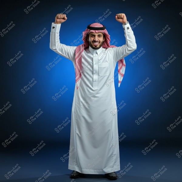 A portrait of an Arab man wearing traditional Saudi white thobe and red ghutra, standing with a smile and raising his fists in a gesture of joy and triumph. The background is dark blue, highlighting his facial features and attire.