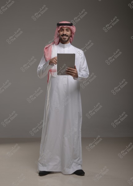 A portrait of a man standing and smiling in a gray studio, wearing traditional white thobe and a red and white checkered keffiyeh. He holds a tablet in his hands, pointing at the screen.