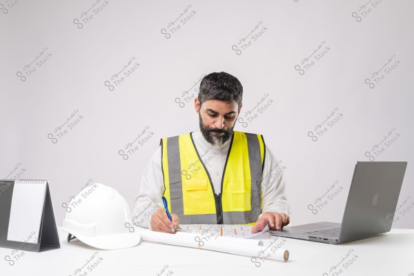 Image of a man sitting at a desk, wearing a traditional Saudi thobe and a yellow safety vest. In front of him is a white helmet and open engineering blueprints on the table, while using a silver laptop on the side. He appears to be reviewing or working on the blueprints.