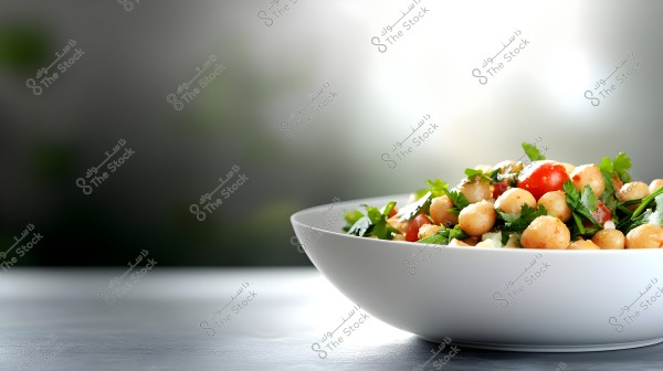 Image of a chickpea salad in a white bowl on a tabletop, featuring chickpeas, tomatoes, and green herbs. The background is blurred with neutral tones.
