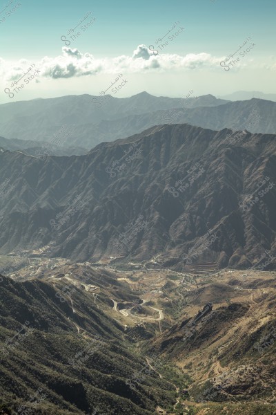 An image of mountainous terrain showing rugged mountain ranges extending to the horizon with some clouds in the clear sky. At the bottom of the image, a winding road is visible passing through the mountains, with some greenery covering the lower areas.